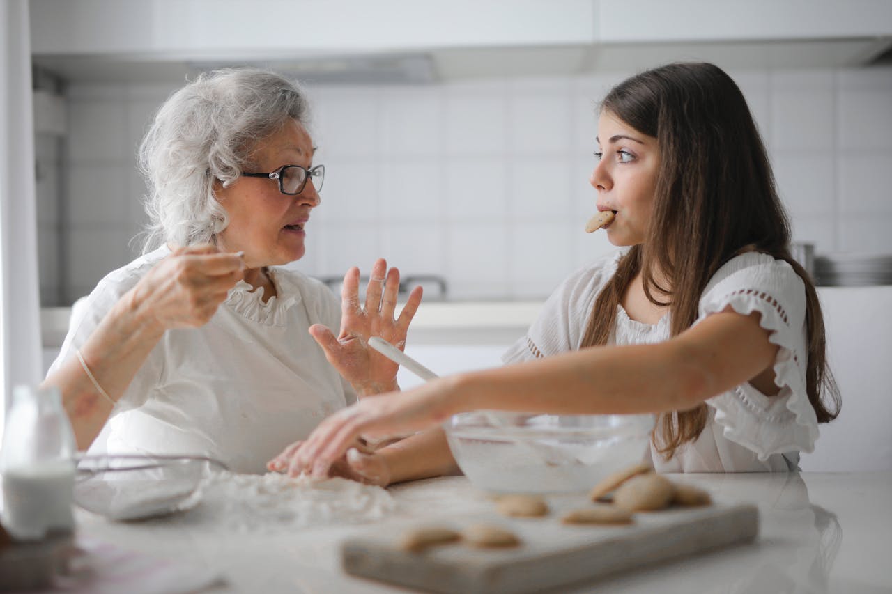 about-us-02 Calm senior woman and teenage girl in casual clothes looking at each other and talking while eating cookies and cooking pastry in contemporary kitchen at home