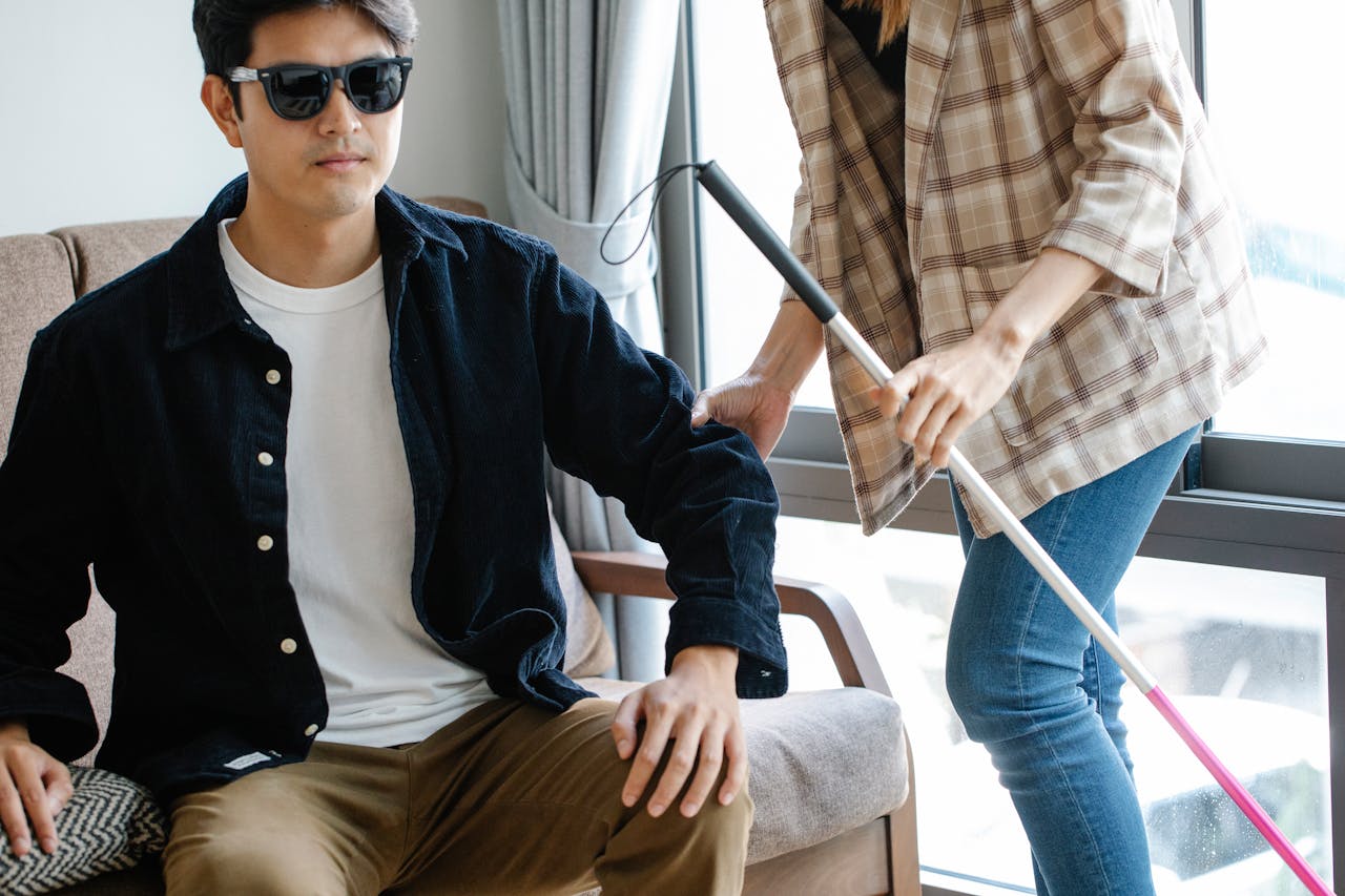 A visually impaired man receives support from a caregiver indoors, using a walking stick.